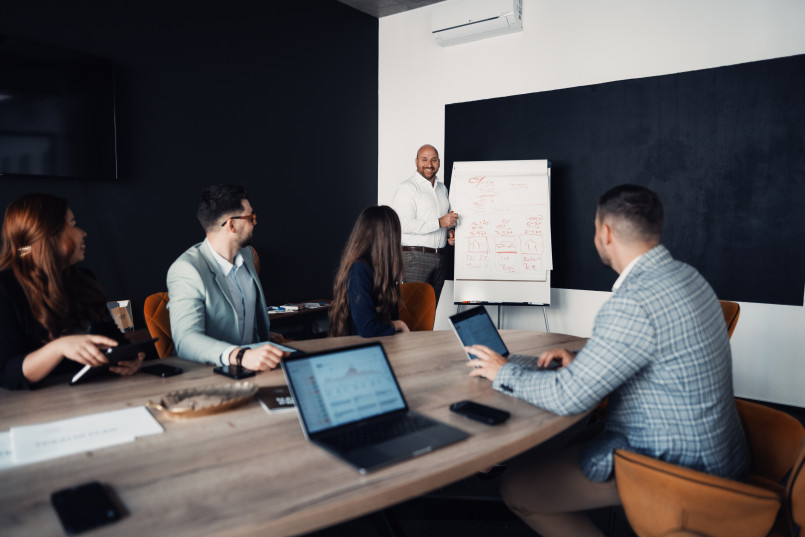 A group of people sitting around a table watching a presentation on a flip chart led by a man standing in front of the board