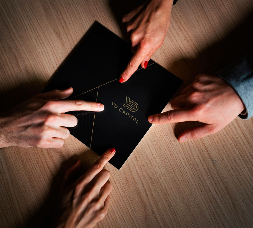 Three hands pointing at a black envelope with the 'YO Capital' logo on a wooden table