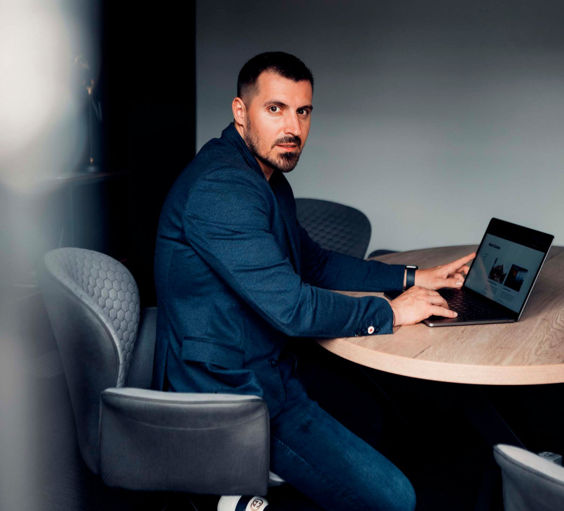 A man in a dark blue blazer sitting at a round table, working on a laptop, looking towards the camera