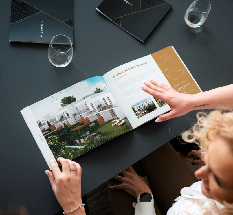 A woman looking through a catalog with photos of modern homes on a black table, with glasses of water and closed envelopes nearby