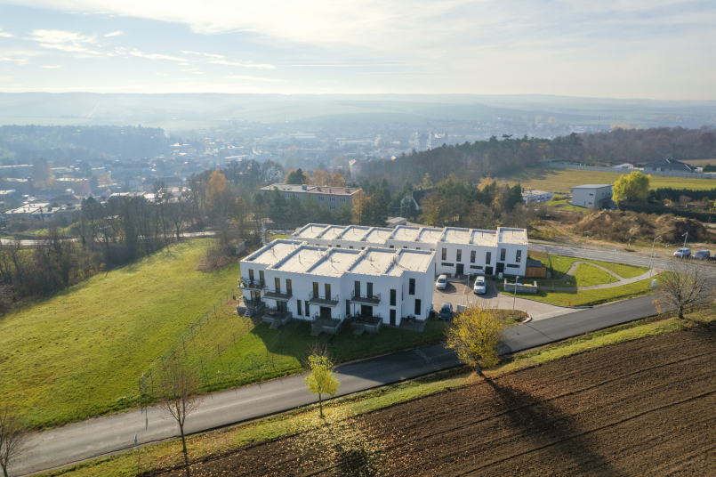 An aerial view of a modern residential complex surrounded by green fields and forests, with a city visible in the background