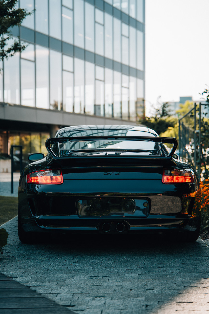 A black Porsche sports car parked on a paved path next to a flower bed, with trees and a modern building in the background.