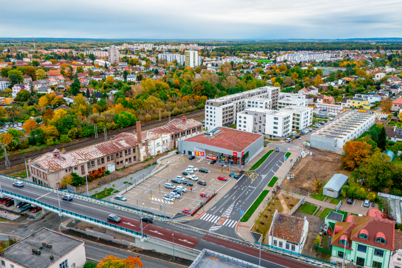 An aerial view of a modern urban area with buildings, roads, and parking lots, surrounded by green trees.