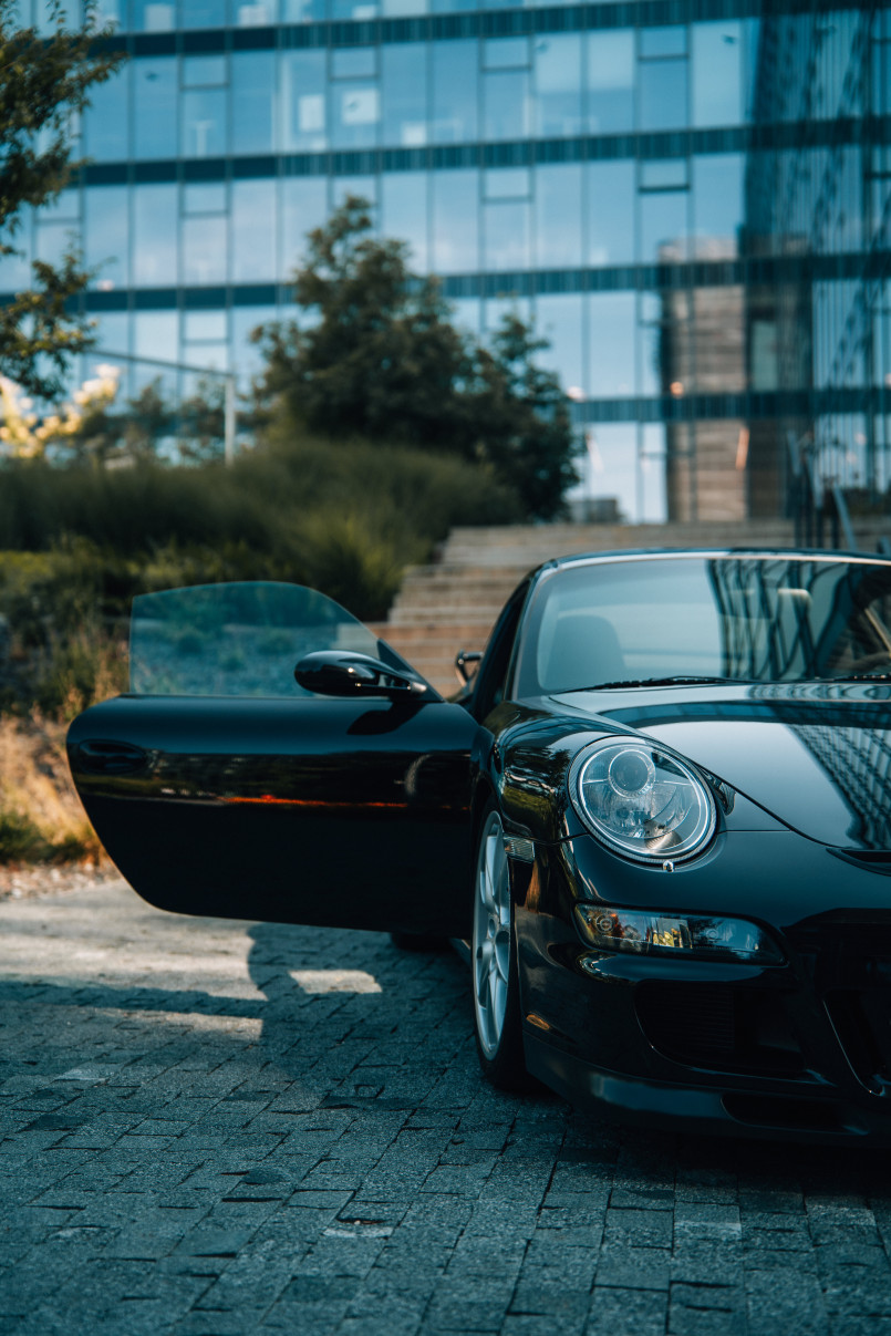 A black Porsche sports car with an open door parked on a paved area, with modern buildings and trees in the background